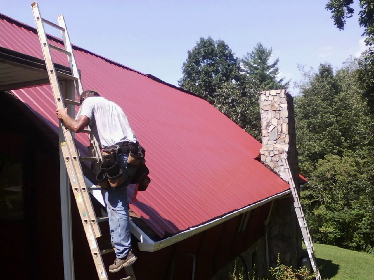 Roofer on a ladder beside a red metal roof and stone chimney — Soffit & Fascia Repair by Roofing Brothers in Elmo